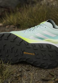 Chaussure de trail bleu clair et verte avec semelle en caoutchouc Continental, montrée par dessous sur une surface extérieure en gravier avec de l'herbe en arrière-plan.