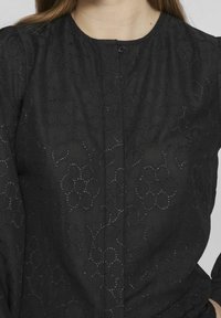 Blusa nera con ricami a occhiello floreali, scollatura rotonda, spalle a palloncino e un singolo bottone nero visibile al centro superiore.