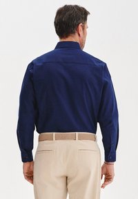 Granatowa koszula z długim rękawem, z guzikami na mankietach, yoke na plecach i gładką teksturą, zestawiona z beżowymi spodniami i plecionym paskiem.