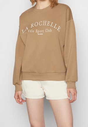 Sweat-shirt beige avec le texte "La Rochelle Vélo Sport Club est. 1998" porté avec un short beige clair sur une personne debout.