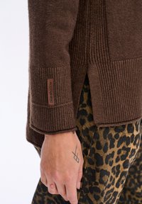 D�łoń z tatuażami geometrycznymi i okręgami obok brązowego swetra z prążkami, oznaczonego hasłem "niebieski cień", oraz spodni z printem w panterkę.