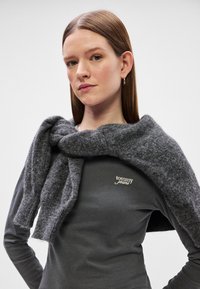 Γκρι μάλλινο κασκόλ κρεμασμένο πάνω σε ένα σκούρο μακρυμάνικο πουκάμισο. Το κασκόλ διαθέτει μια απαλή, υφασμάτινη υφή με έναν κόμπο μπροστά.