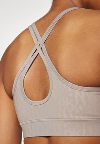 Light taupe sports bra with a textured design, featuring crossed straps and an open back. DKNY logo in subtle blue at the band.