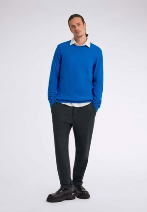 Blauer Langarmpullover über einem weißen Kragenhemd, kombiniert mit dunkelgrauen Hosen und schwarzen Plateauschuhen. Einfaches, gerade geschnittenes Design.