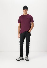 T-shirt en coton bordeaux avec un petit logo jaune, associée à un jean noir et des chaussures de sport argentées. Le modèle porte une casquette noire.