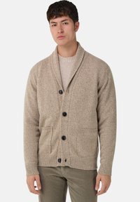 Ung man som bär en beige sjalkragekofta med svarta knappar, två framlommer och bruna byxor, stående mot en vit bakgrund.