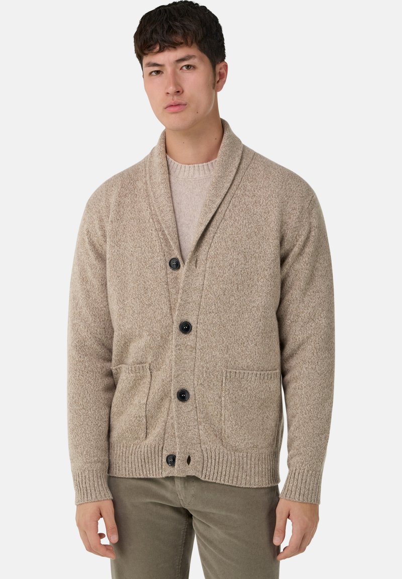 Ung man som bär en beige sjalkragekofta med svarta knappar, två framlommer och bruna byxor, stående mot en vit bakgrund.