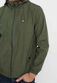 Homme portant une veste à capuche zippée olive avec un logo d'oiseau jaune sur la poitrine gauche et les mains dans les poches.
