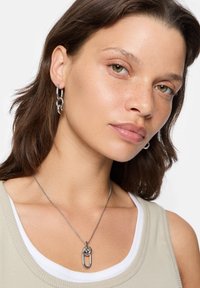 Metallaccessoarer inkluderar ett silverfärgat kedjehalsband med en stor, sammanflätad oval länk och en matchande hoop-örhänge, placerat mot en ljusbeige överdel.