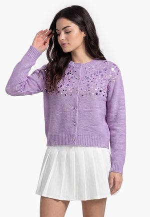 Donna con lunghi capelli scuri che indossa un cardigan lavanda con paillettes e bottoni a forma di cuore, abbinato a una gonna plissettata bianca.