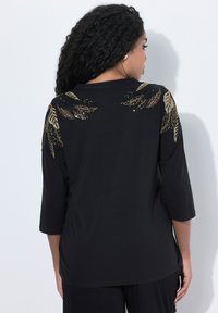 Maglietta nera a tre quarti con motivi a foglia in paillettes dorate e nere sulle spalle. Materiale morbido con una vestibilità rilassata.