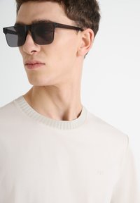 Gafas de sol negras rectangulares con lentes oscuros, llevadas con una camiseta de cuello redondo beige claro con un collar acanalado y un sutil logotipo bordado.