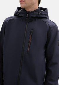 Marineblaue wasserdichte Jacke mit Kapuze, Frontreißverschluss, seitlichen Taschen mit roten Akzenten und strukturiertem Stoff. Verfügt über verstellbare Zugbänder.