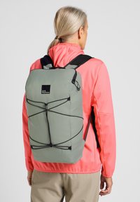 Groene lichtgewicht rugzak met een zwart logo, voorzien van elastische koorden voor aanpassing en gevoerde schouderbanden. Draag door een persoon in een roze jas.