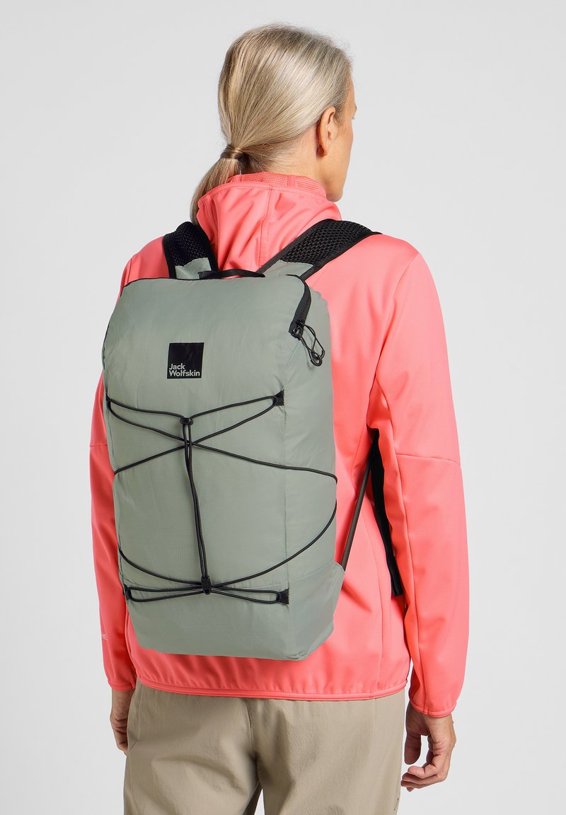 Groene lichtgewicht rugzak met een zwart logo, voorzien van elastische koorden voor aanpassing en gevoerde schouderbanden. Draag door een persoon in een roze jas.