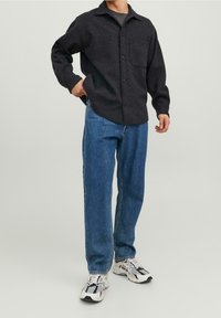 Camicia nera a bottoni con taschino sul petto, abbinata a jeans blu a gamba dritta e sneaker bianche con dettagli neri. Sfondo neutro.