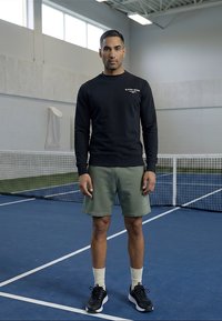 Mies, jolla on musta collegepaita, vihreät shortsit ja mustat lenkkarit, seisoo sinisellä sisätenniskentällä verkon lähellä.