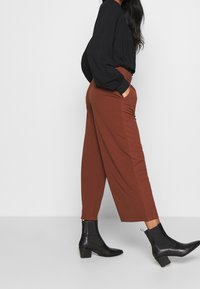 Hnědé široké kalhoty s bočními kapsami, doplněné černou halenkou s dlouhými rukávy a černými kotníkovými botami s blokovým podpatkem.