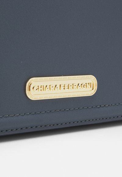 Borsa in ecopelle blu navy con una placca del logo placcata in oro; presenta cuciture dettagliate lungo i bordi e una superficie liscia e testurizzata.