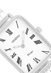 Montre carrée en argent avec des chiffres romains sur un cadran blanc, bracelet en acier inoxydable et couronne d'ajustement, affichant la marque "oozoo".