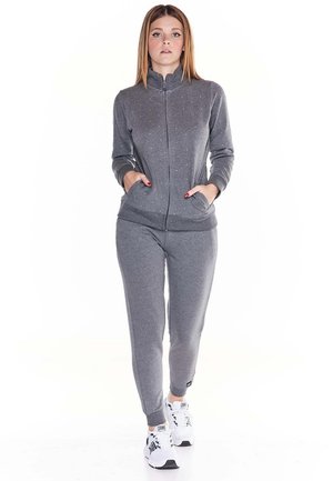 Une femme présente un survêtement gris zippé, avec un pantalon de jogging assorti et des baskets blanches, sur un fond blanc uni.