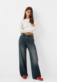 Široke denim hlače temnega pranja s visokim pasom in rahlo obrabljeno teksturo. V kombinaciji z oprijetim belim dolgim rokavom in rdečimi čevlji.