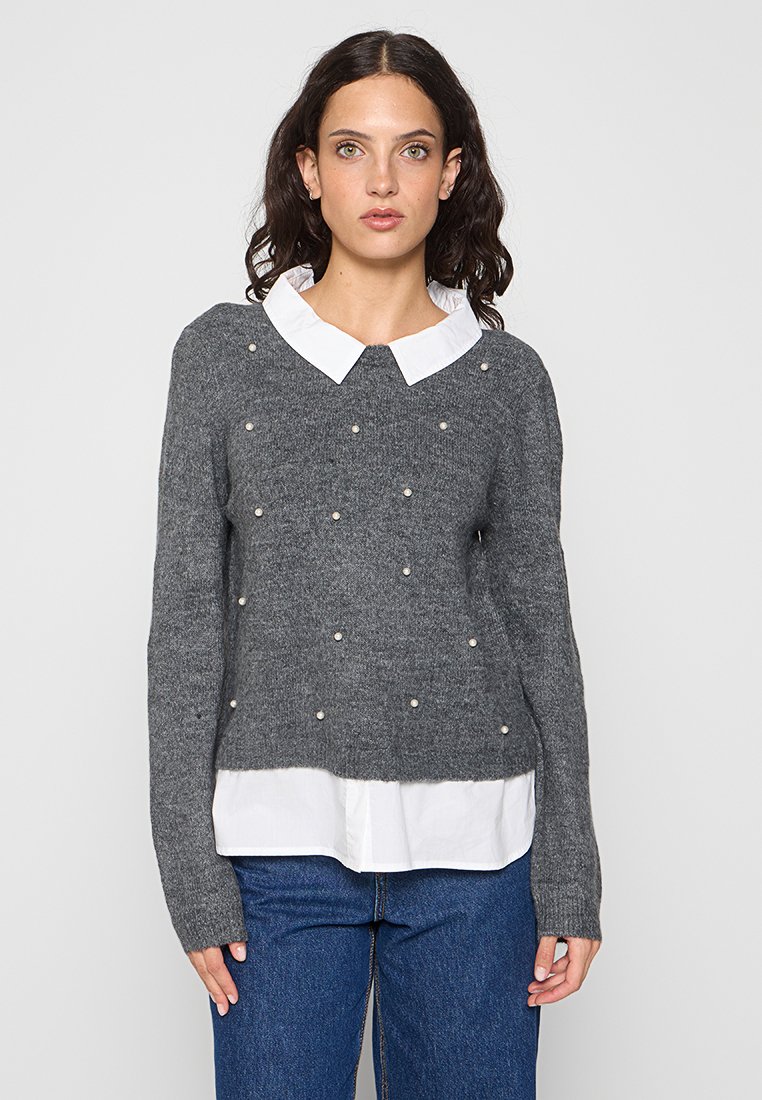 Grauer Strickpullover mit rundem Ausschnitt, verziert mit weißen Perlen und über einem weißen Hemd mit Kragen getragen. Lockerer Schnitt und lange Ärmel.