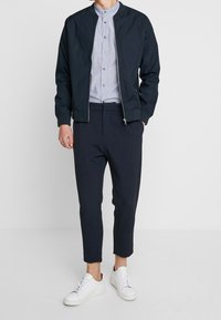 Marinblå bomberjacka över en ljusblå button-up skjorta med ståkrage, tillsammans med mörka tapered byxor och vita sneakers. Släta texturer genomgående.