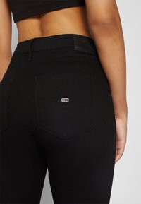 Lähikuvassa on inimene, kes kannab kõrge vöökohtaga musti teksaseid, mille paremas tagataskus on väike ristkülikukujuline logo, ja musta kropp-topi.
