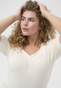 Haut ajusté crème avec un décolleté en V et un détail froncé au centre. Manches courtes et texture douce et lisse.