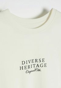 Kremowa bawełniana koszulka z okrągłym dekoltem, na której w czarnym kolorze wydrukowane jest "DIVERSE HERITAGE" oraz "OriginalEth". Gładka faktura, casualowy design.