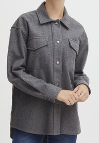 Szara koszula-kurtka z mieszanki wełny, z kołnierzem, zatrzaskami i dwoma kieszeniami na piersi. Posiada luźny krój i długie rękawy.