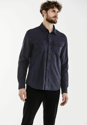 Camisa de botones oscura de color marino, hecha de una tela suave, con dos bolsillos en el pecho y un dobladillo curvado. Las mangas largas están enrolladas en los puños.