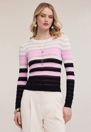 Mujer con suéter de rayas acanalado de manga larga en blanco, rosa y negro, combinado con pantalones altos de color crema y joyería dorada.