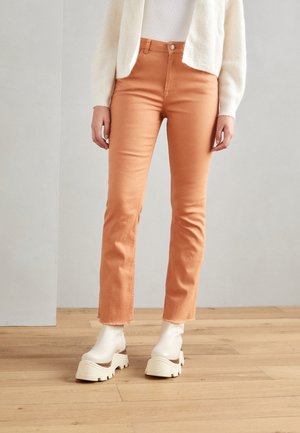 Persoon draagt oranje broek met hoge taille en recht gesneden pijpen met rafelige zomen, gecombineerd met witte chunky platform enkellaarsjes, staande op een houten vloer.
