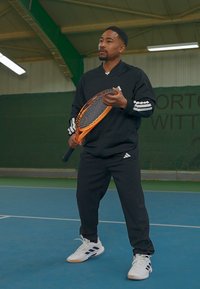 Black athletic tracksuit with white accents, holding an orange tennis racket, on a blue indoor court with green walls.