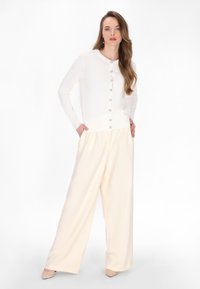 Cardigan bianco a maniche lunghe con scollatura rotonda e chiusura con bottoni, abbinato a pantaloni larghi color crema e tacchi nudi. Texture liscia.