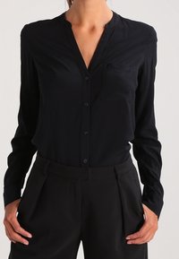 Chemise noire à boutons, avec un col classique, des manches longues et une poche poitrine gauche. Confectionnée en tissu lisse, elle présente un design minimaliste.