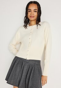 Cardigã de malha creme com decote redondo, fecho de Botão e bainha canelada. Combinado com uma saia plissada cinza riscada.