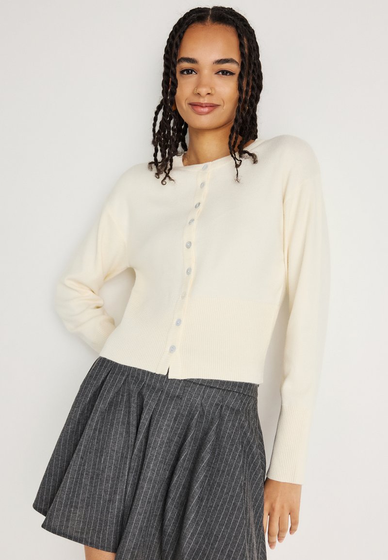 Cardigã de malha creme com decote redondo, fecho de Botão e bainha canelada. Combinado com uma saia plissada cinza riscada.