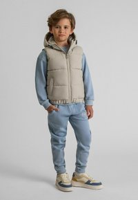 Ljusbeige puffväst med huva, grå dragkedja; blå bomullströja, matchande fleecbyxor och beiga sneakers med blå och guldakcent.