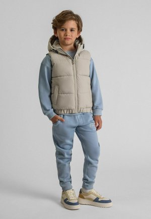 Gilet rembourré beige clair avec capuche, fermeture éclair grise ; sweat-shirt en coton bleu, pantalon en polaire assorti et baskets beiges avec accents bleus et dorés.