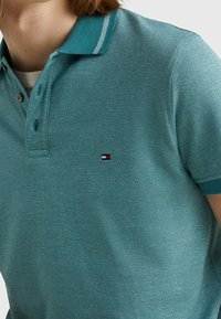 Teal polo tričko s krátkým rukávem, bílým pruhem na límci a malým vyšitým červeným, bílým a černým obdélníkovým logem na hrudi.