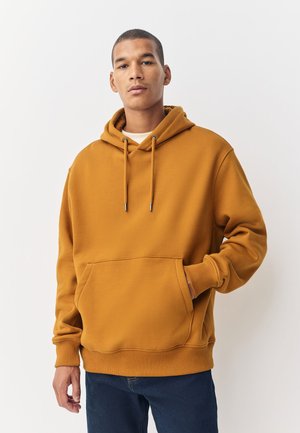 Jeune homme portant un sweat à capuche couleur moutarde avec poche avant et cordons, une main dans la poche, sur un fond blanc uni.