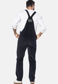 Marineblaue Corduroy-Latzhose mit verstellbaren Trägern, einer Gesäßtasche und einem Etikett. Der Stoff hat eine weiche, strukturierte Oberfläche und eine entspannte Passform.