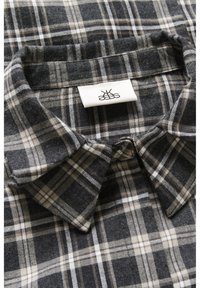 Cămașă din flanel cu un model de carouri în nuanțe de negru, gri și alb. Are un guler ascuțit și o etichetă țesută la gât. Textură moale și material cald.