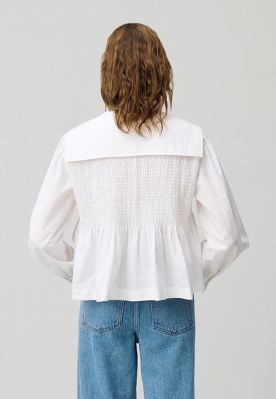 Blusa de algodón blanca con la espalda fruncida, que presenta un cuello amplio y mangas abullonadas. Combinada con jeans de mezclilla azul de cintura alta.