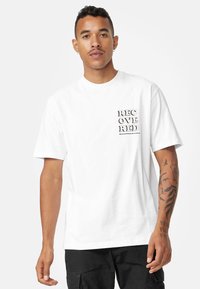 Hvid bomulds-T-shirt med sort trykt tekst, der siger "REC OVER RED" på den øverste venstre side. Modellen står med afslappede arme og bærer sorte bukser.