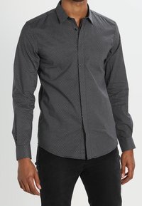 Czarna, długiego rękawka koszula z delikatnym białym wzorem w kropki, z kołnierzykiem, zapięciem na guziki i dopasowanym krojem. Noszona z czarnymi spodniami.