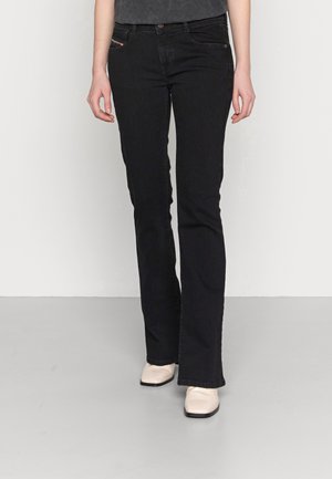 Osoba ubrana w czarne jeansy o prostych nogawkach i białe buty w wydłużonym kształcie, stojąca na szarej podłodze na tle jednolitej białej ściany.